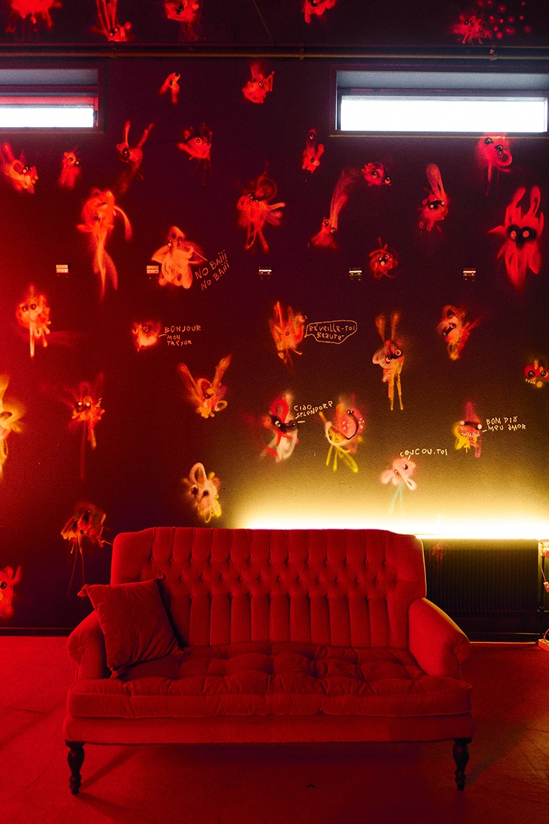 Rood verlichte lounge met een gecapitonneerde bank voor een muur vol neon schepsels van Simonian bij Dutch Art Room in Rotterdam